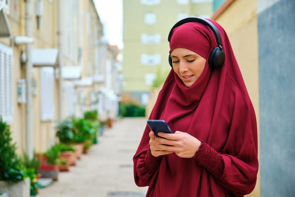 Muslim young woman in hijab using phone and headphones in the street.